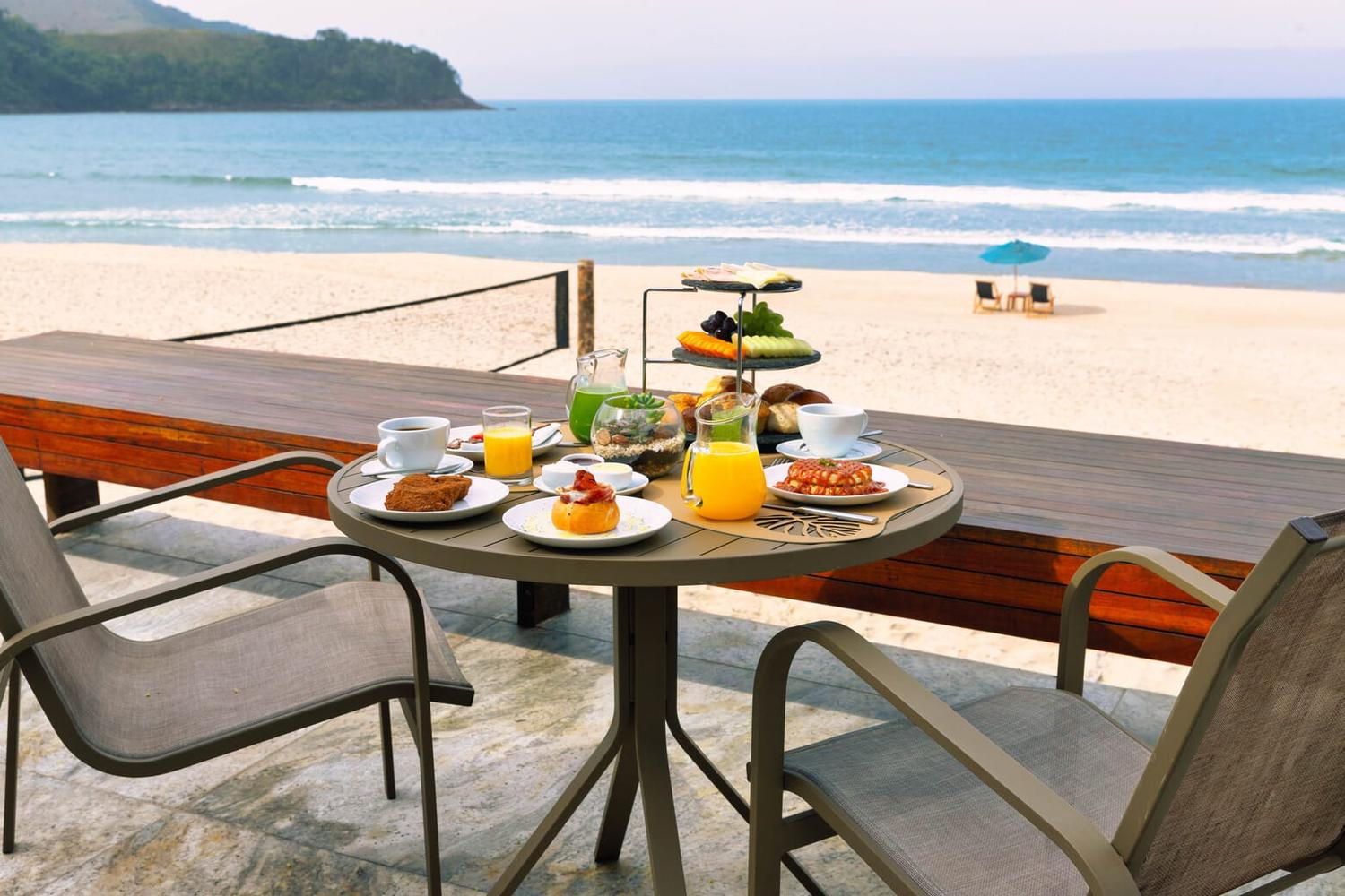 Mesa de café da manhã com frutas, sucos e xícaras, diante da praia de Maresias.