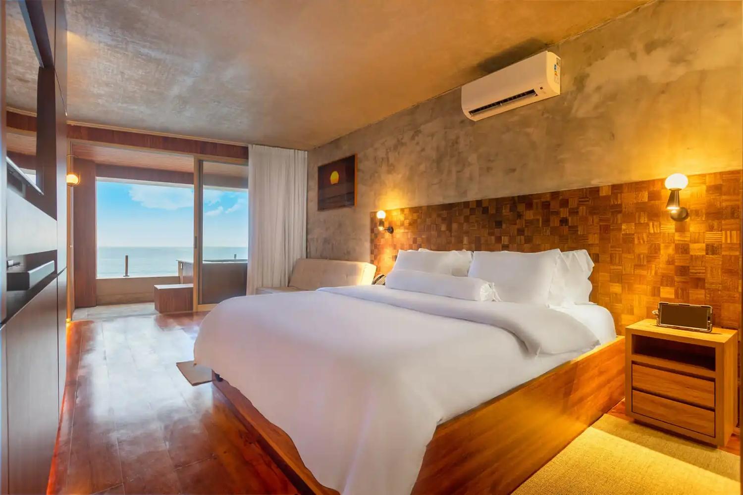 Acomodação com cama de casal, cabeceira de madeira e varanda voltada para o mar.