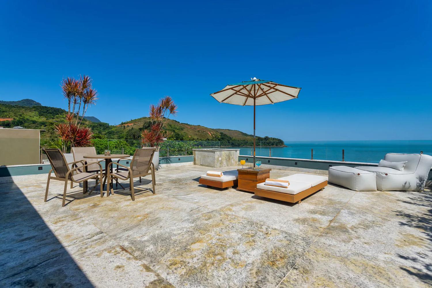 Terraço com espreguiçadeiras, guarda-sol e vista aberta para o oceano e a costa.